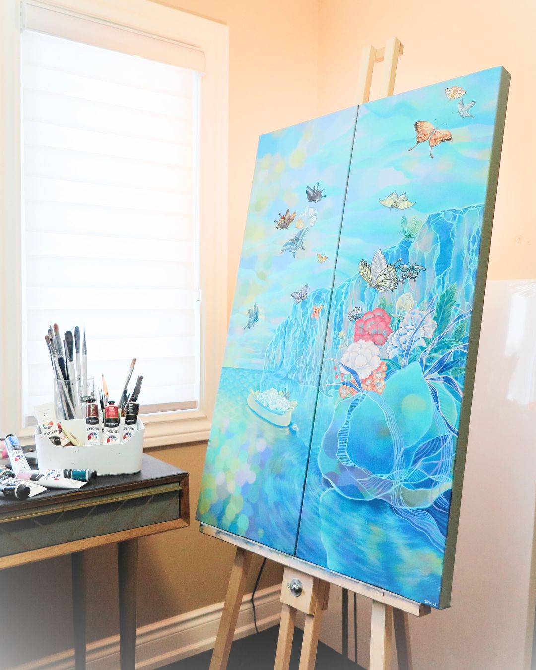 A painting of a rock cliff, flowers, butterflies and the lake on an art easel. 