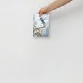 Hand holding a 5x7 inch art print with a bluejay and tree design on a light gray background, which symbolizes freedom and prosperity.