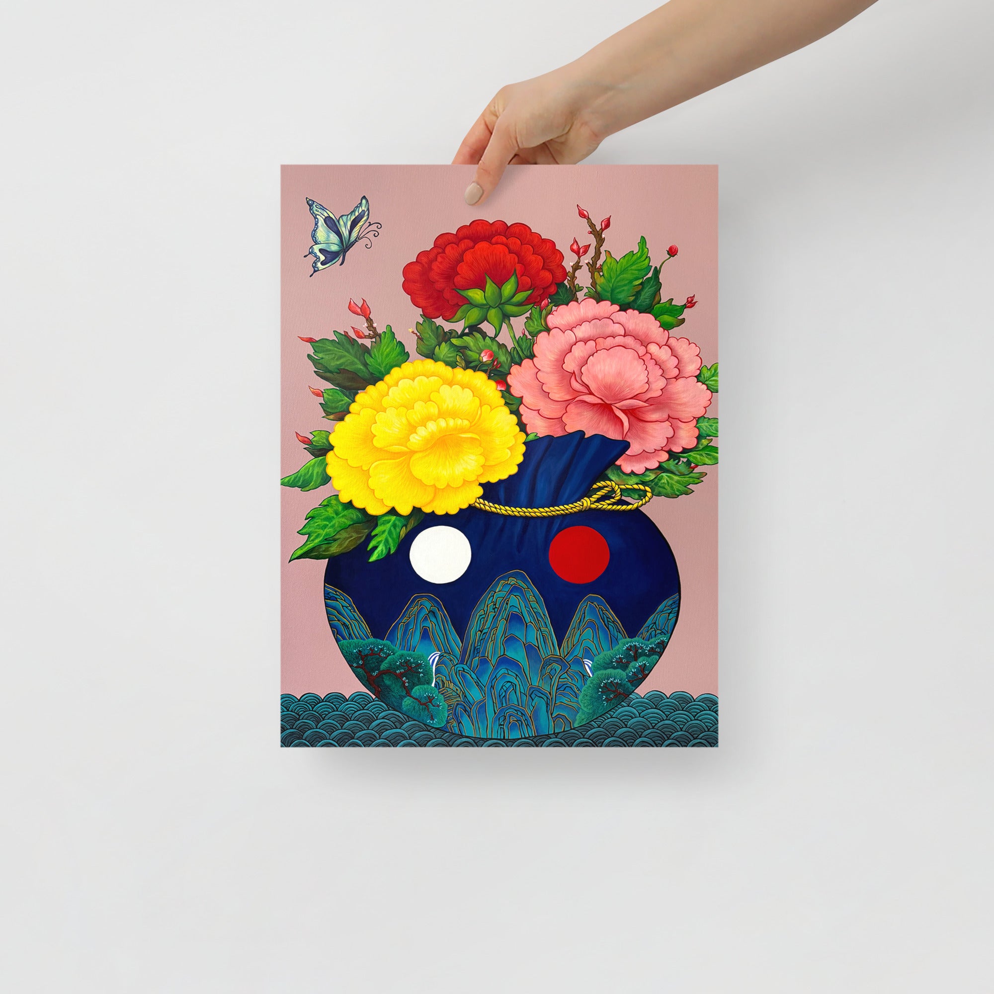Hand holding a 12x16 inch art print of a Korean coih pouch adorned with traditional motifs inspired by Minhwa art holding a bouquet of colorful peonies with a butterfly floating in the corner, representing good fortune, harmony and joy, on a white background.
