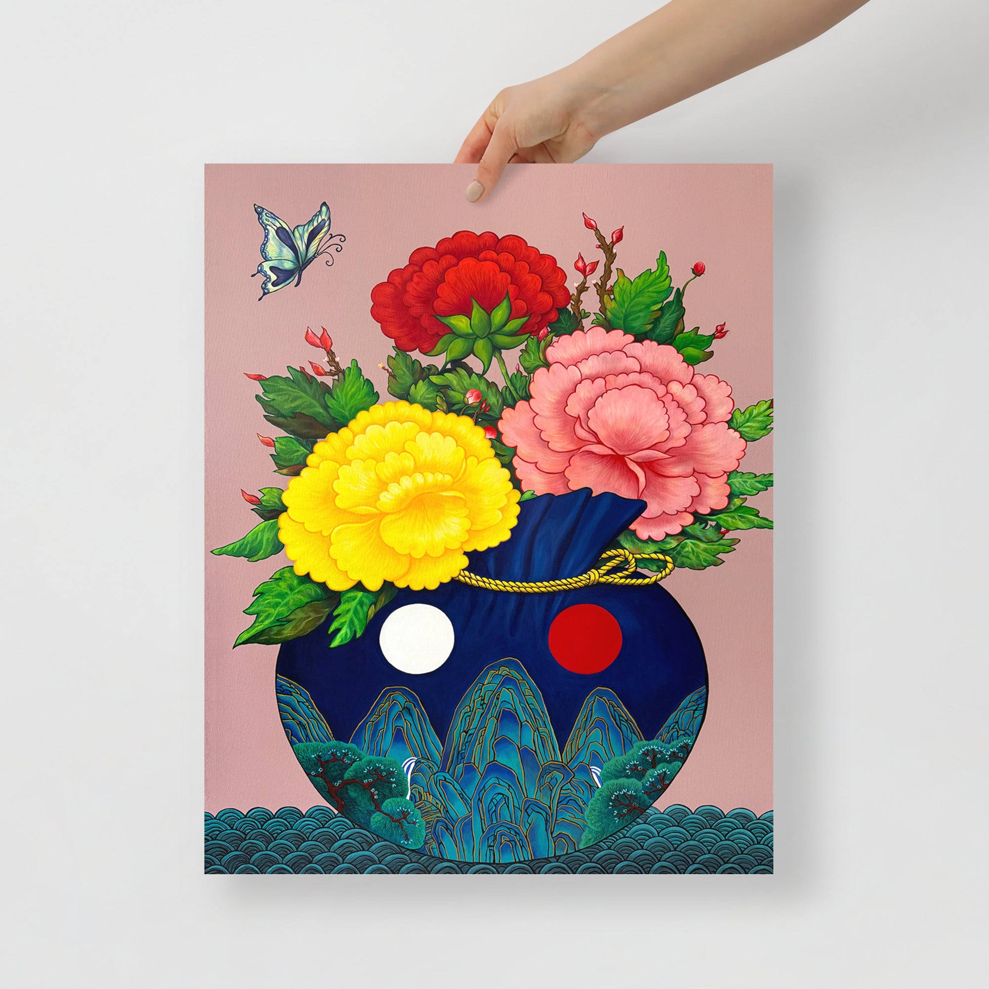 Hand holding a 16x20 inch art print of a Korean coih pouch adorned with traditional motifs inspired by Minhwa art holding a bouquet of colorful peonies with a butterfly floating in the corner, representing good fortune, harmony and joy, on a white background.