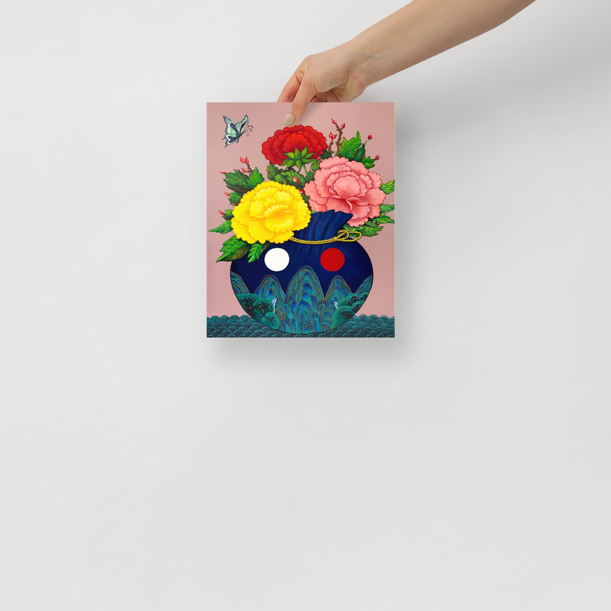 Hand holding a 8x10 inch art print of a bokjumeoni adorned with traditional motifs inspired by Minhwa art holding a bouquet of colorful peonies with a butterfly floating in the corner, representing good fortune, harmony and joy, on a white background.