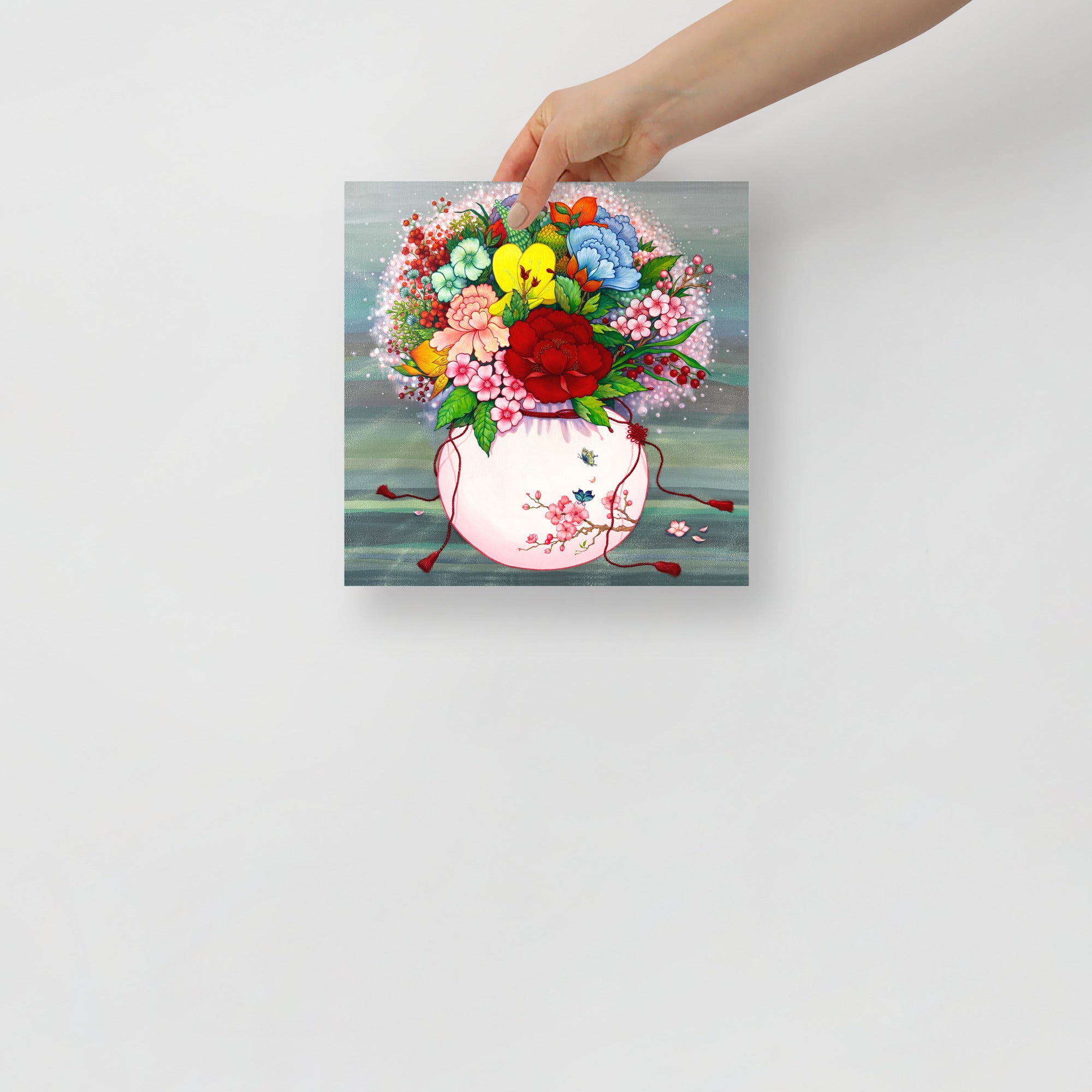 Hand holding a 10x10 inch art print that draws from Korean folk art heritage, weaving together blossoms and traditional motifs that celebrate harmony, wellness, and good fortune, on a light gray background.