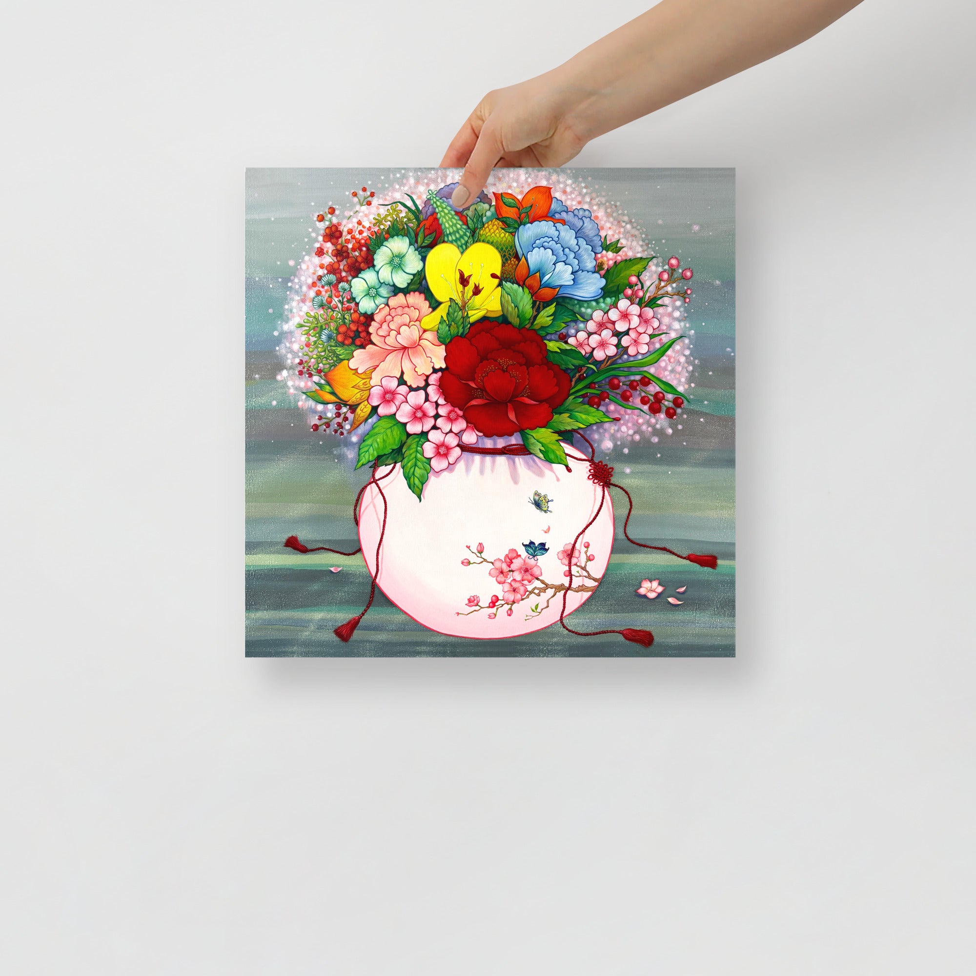 Hand holding a 14x14 inch square art print that draws from Korean folk art heritage, weaving together blossoms and traditional motifs that celebrate harmony, wellness, and good fortune.
