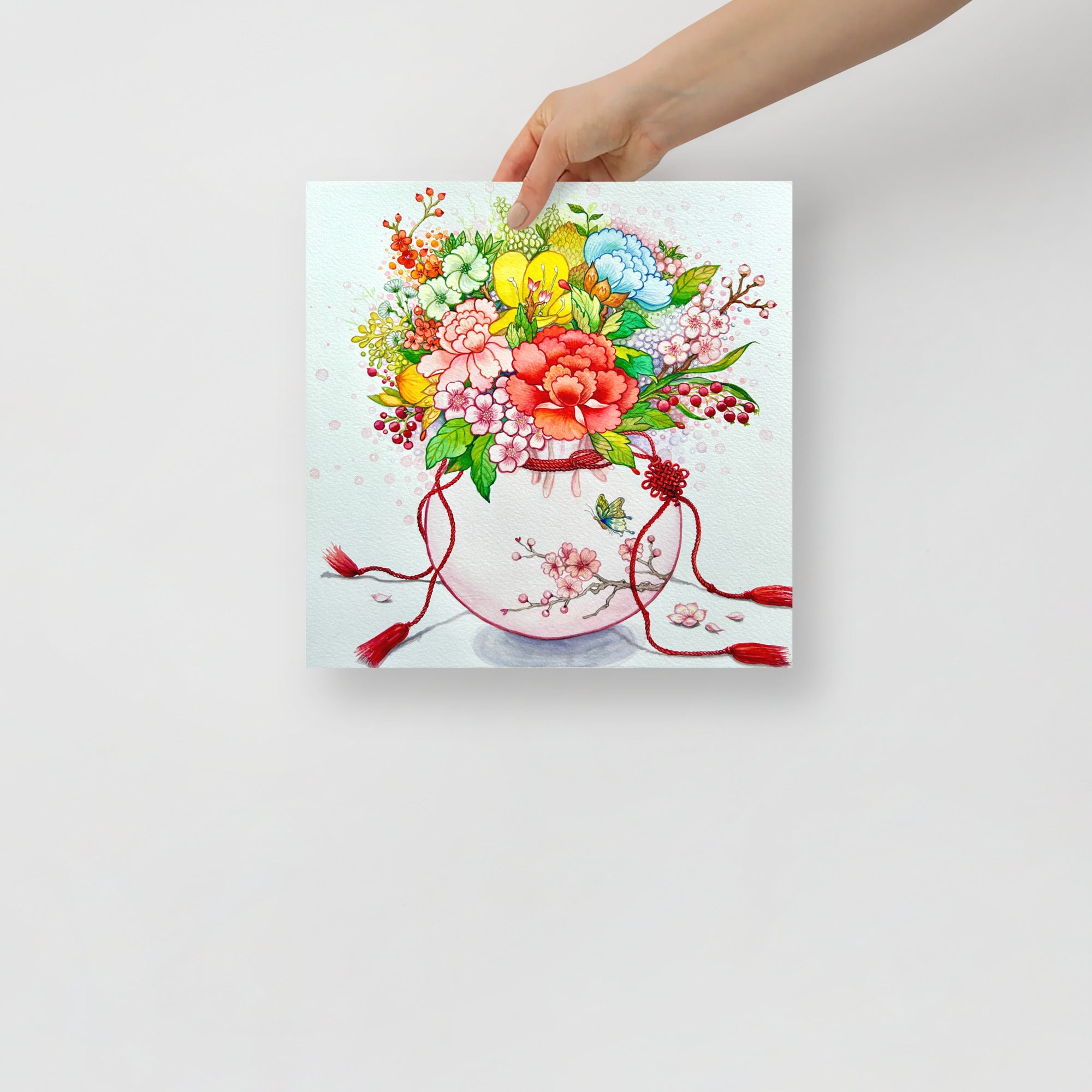 Hand holding a square canvas print of a colorful floral design with red ribbons on a light gray background (12x12 size)